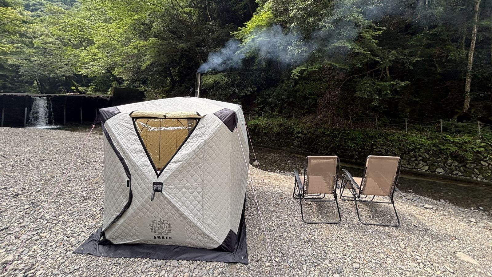 川辺に設営したAMBERテントサウナ。煙突から煙が上がり、奥に滝、手前に外気浴用のチェア2脚